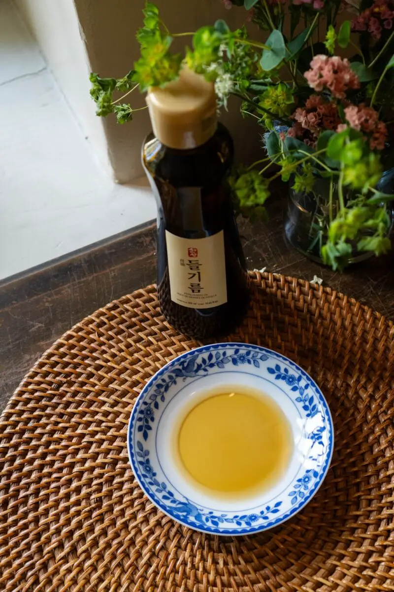 A photograph of deulgireum on a blue and white curved plate. The perilla oil bottle sits in the background. It sits on  a wicker place mat. 