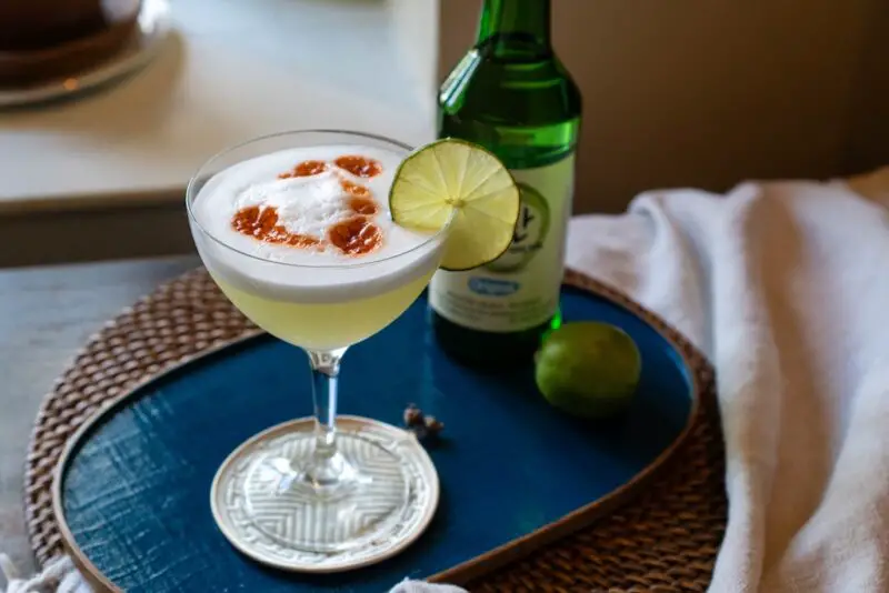 A side shot of this soju sour in a glass. Bitters sit on top of the egg foam. A lime sits on the side of the glass. A bottle of soju is in the background. Everything sits on a blue tray. 