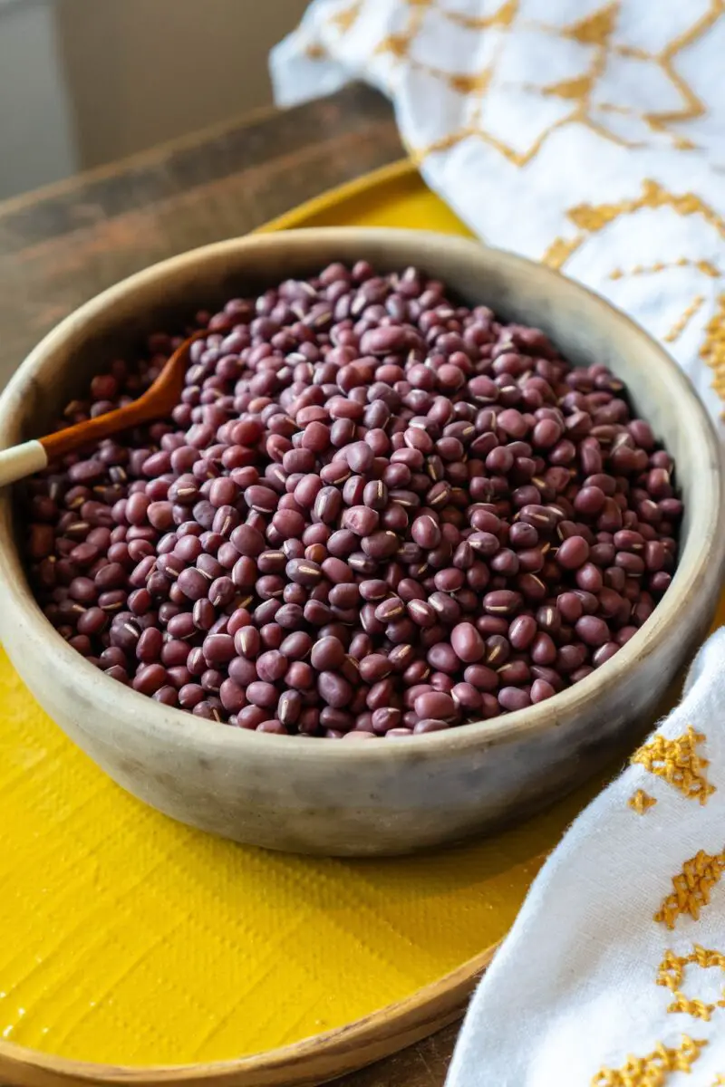 A closeup photo of dried red adzuki beans in a bowl. The bowl is sitting on a yellow tray next to a table cloth. 