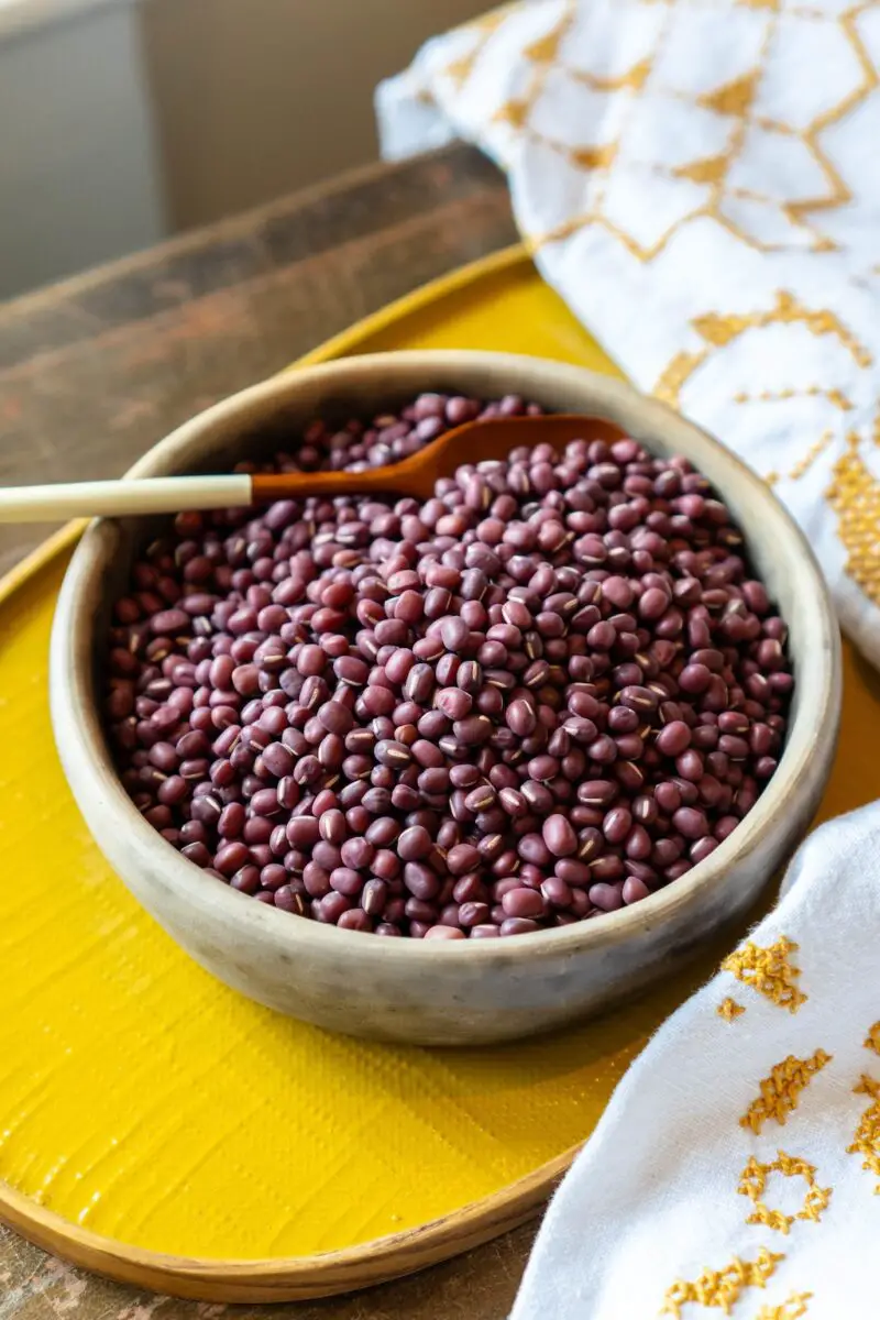 A photo of dried red adzuki beans in a bowl. The bowl is sitting on a yellow tray next to a table cloth. 