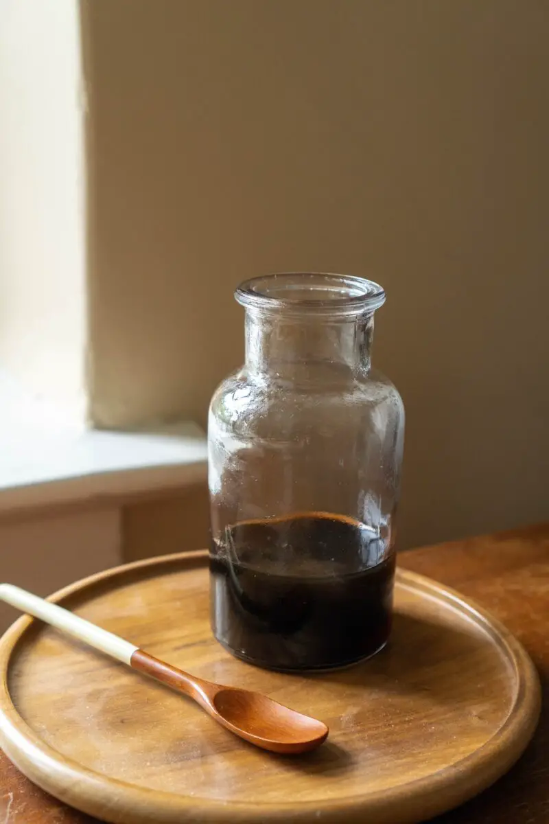 A photo of burnt sugar syrup in a glass container. The container sits on a wooden tray. A wooden spoon sits next to it. 