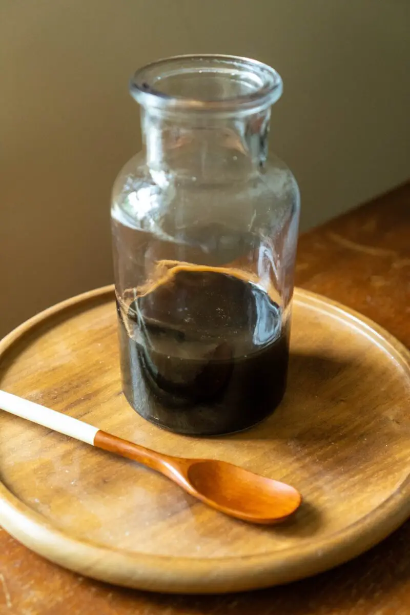 A photo of burnt sugar syrup in a glass container. The container sits on a wooden tray. A wooden spoon sits next to it. 
