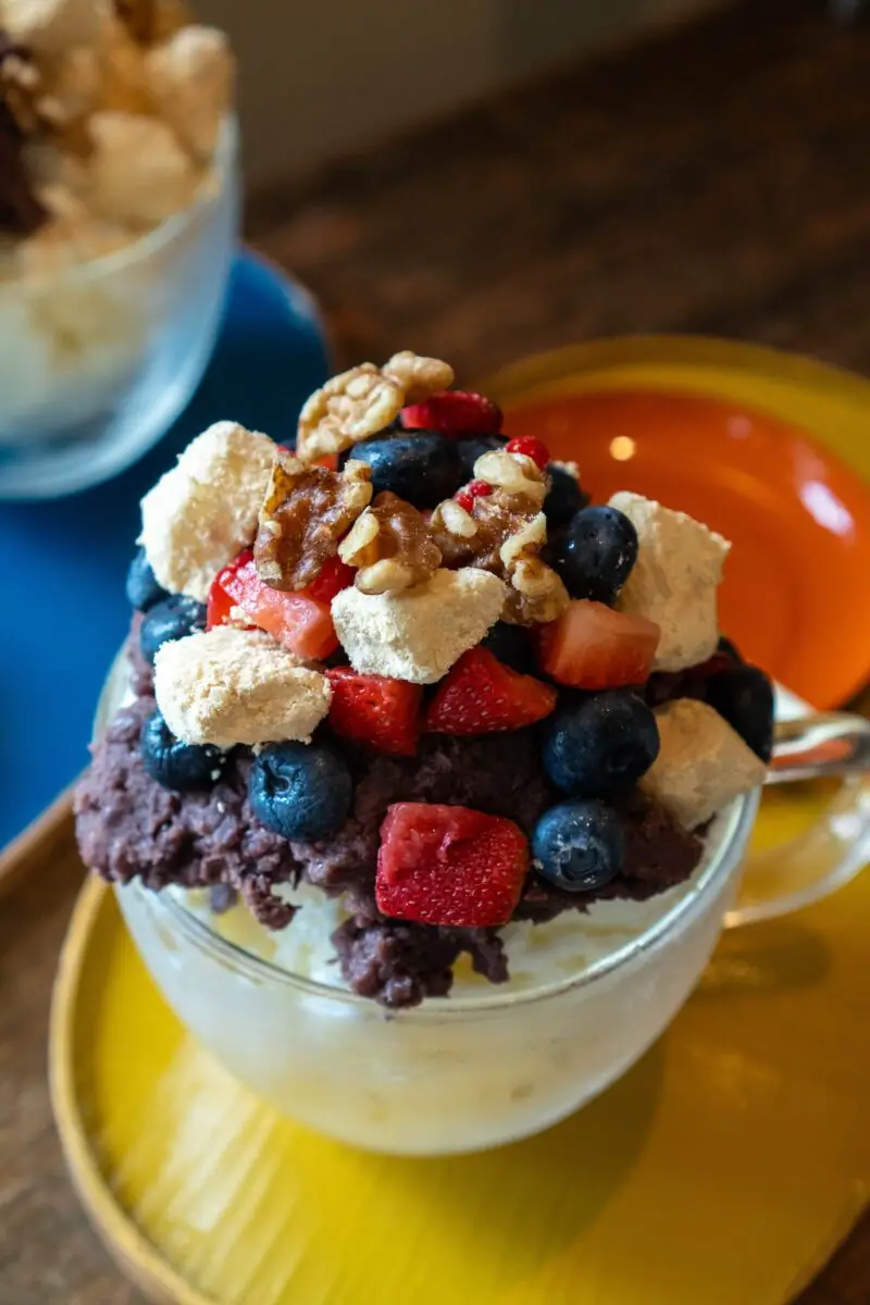 A photo of red bean shaved ice in a clear bowl on a yellow tray. Spoons sit on a orange plate on the tray. The shaved ice is topped with sweet red bean paste, fruit, injeolmi, and nuts.
