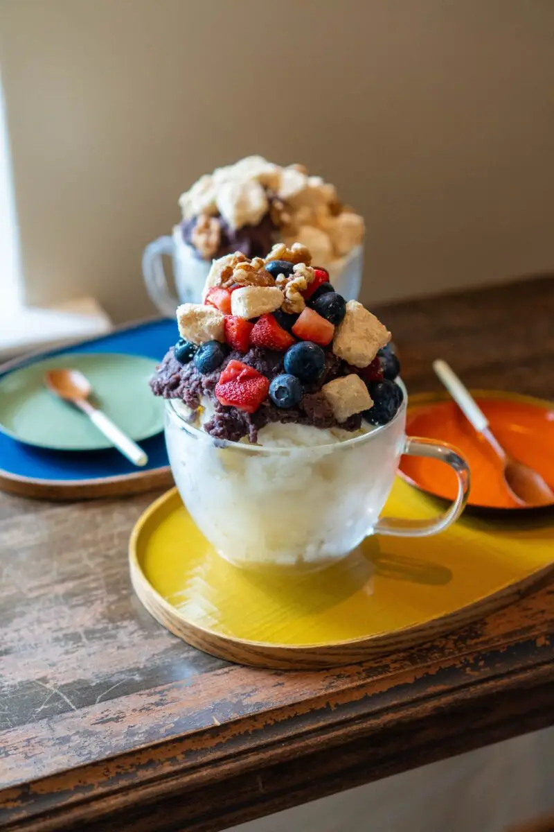 A photo of two types of milk shaved ice. Sweet red bean shaved ice sits in the front on a yellow tray. It is topped with sweetened red beans, fruit, nuts, and injeolmi. Injeolmi shaved ice sits in the background on a blue tray.