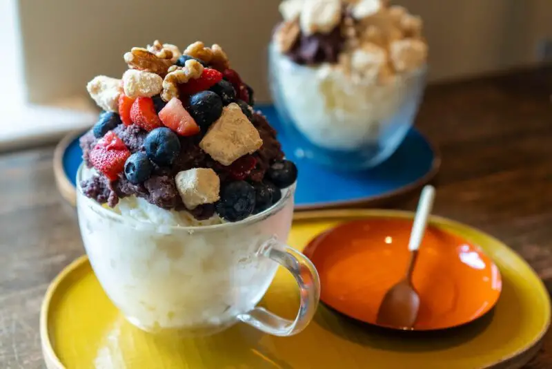 A photo of Korean patbingsu in a clear bowl on a yellow tray. Spoons sit on a orange plate on the tray. The bingsu is topped with Korean sweet red bean paste, fruit, injeolmi, and nuts. Injeolmi bingsu sits on the background on a blue tray.