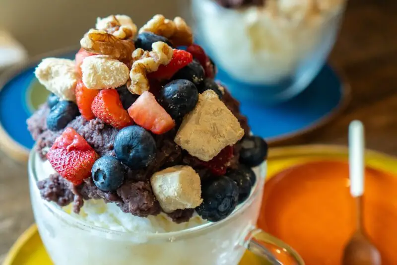 A photo of Korean patbingsu in a clear bowl on a yellow tray. Spoons sit on a orange plate on the tray. The bingsu is topped with Korean sweet red bean paste, fruit, injeolmi, and nuts. Injeomi bingsu sits in the background on a blue tray.